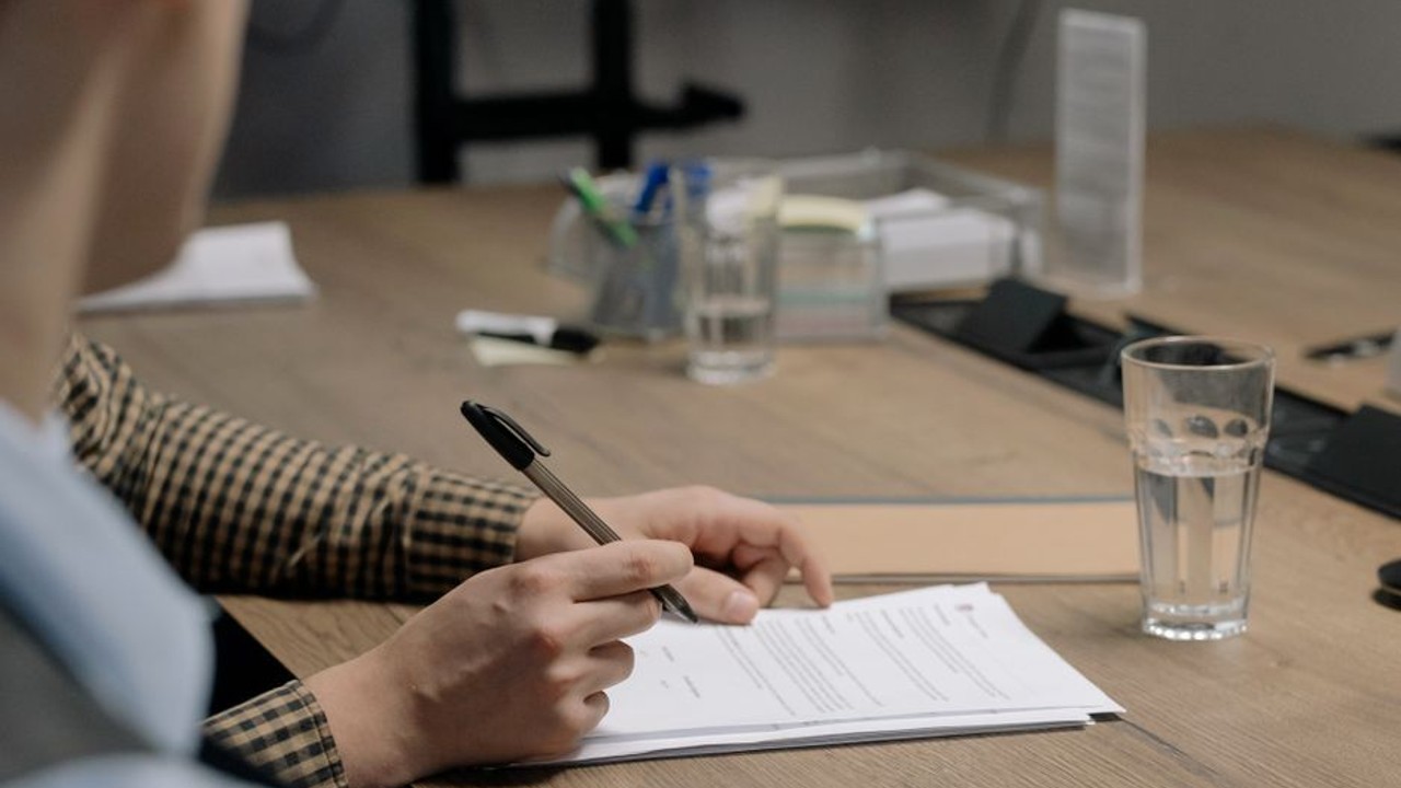Close-up of a person's hands reviewing car title documents and a bill of sale on a desk, with a pen ready to sign. The documents indicate vehicle ownership transfer.