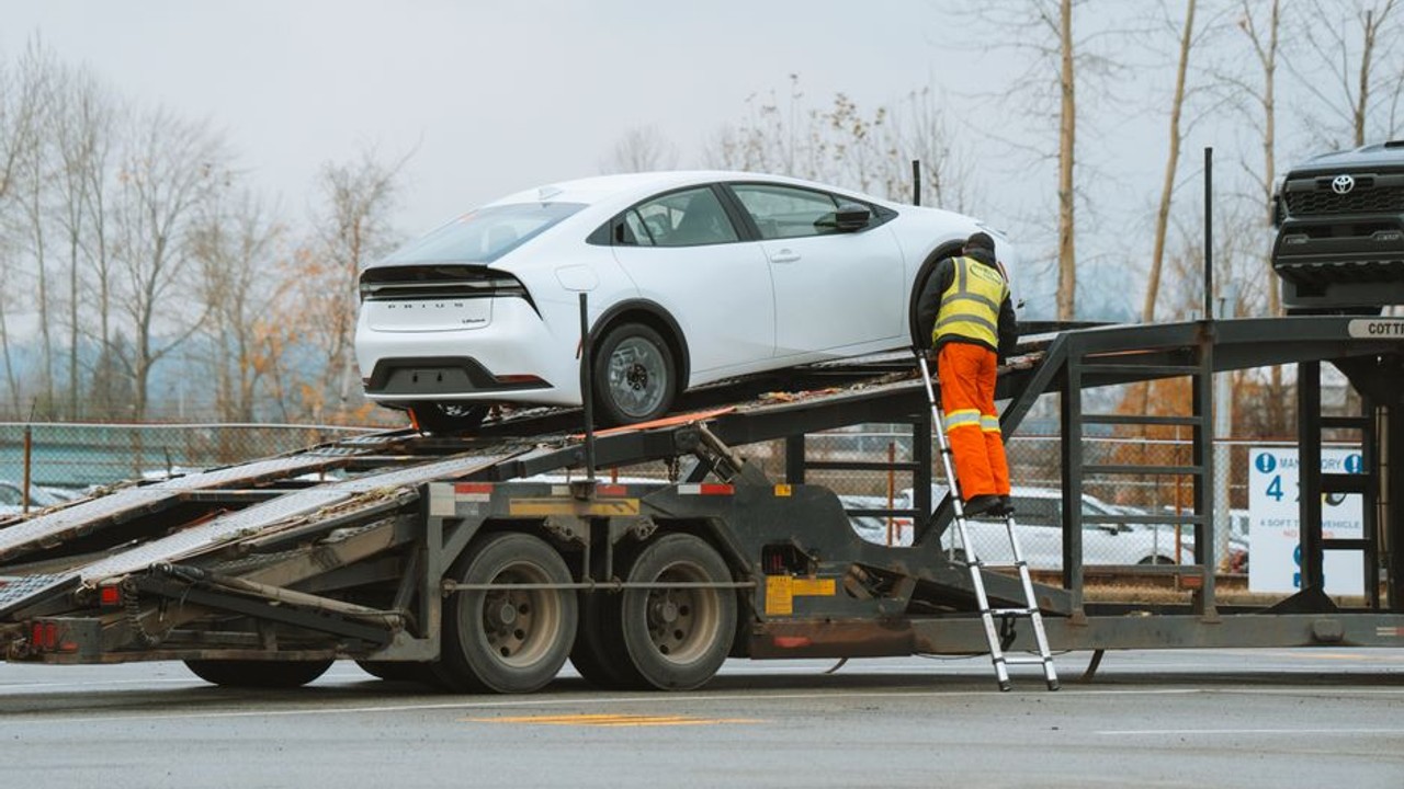 Professional car carrier truck equipped with a winch, carefully loading a damaged, non-running sedan onto its upper deck at an auction lot. The driver is overseeing the process.