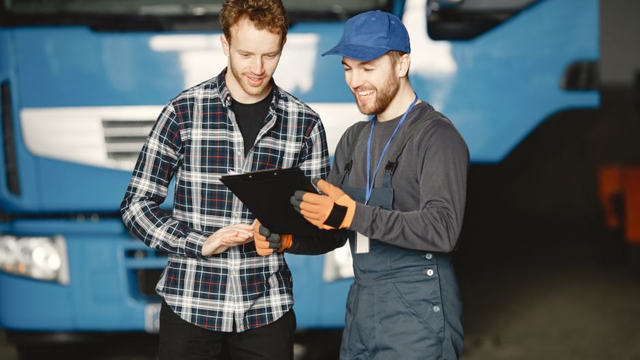 Two professional car carrier drivers are having a discussion with an auction house employee at a vehicle release gate, holding clipboards and reviewing paperwork. Large trucks are visible in the background.