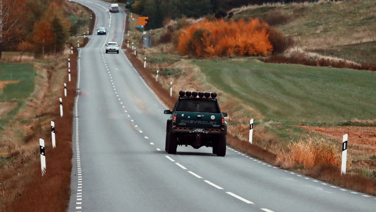 Modern car carrier truck driving on a winding highway through a forest with vibrant fall foliage, clear autumn sky.