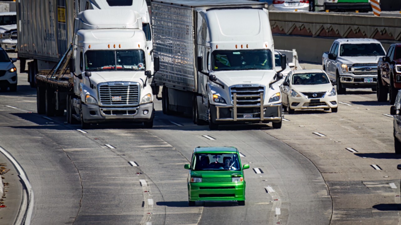Open car carrier truck transporting various sedans and SUVs on a busy multi-lane highway under a blue sky, demonstrating economical vehicle transport