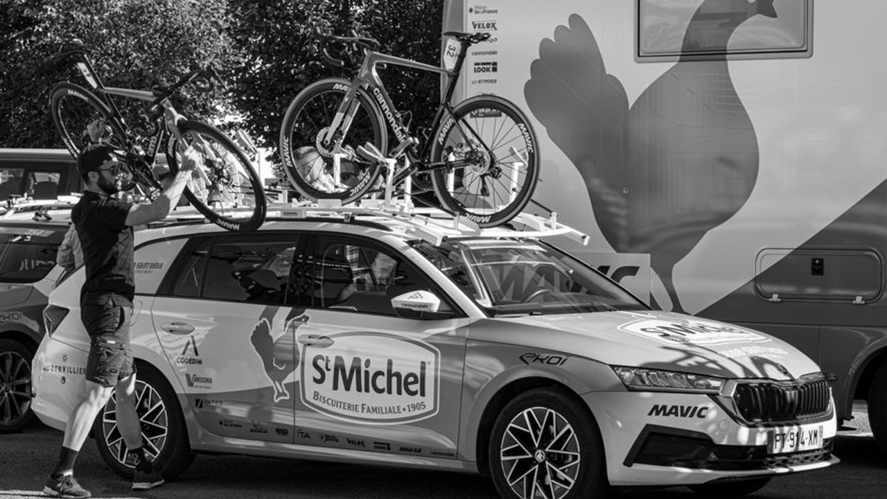 Car with a large roof rack and bike mounts being prepared for loading onto an open car carrier, with a transport specialist inspecting the setup.